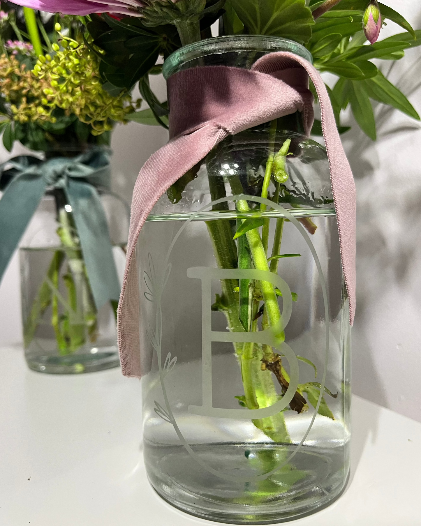 Personalised apothecary-shaped flower vase with the letter B engraved in a delicate oval border, filled with water and flower stems. Another vase is visible out of focus in the distance, highlighting the personalised apothecary vases available for sale