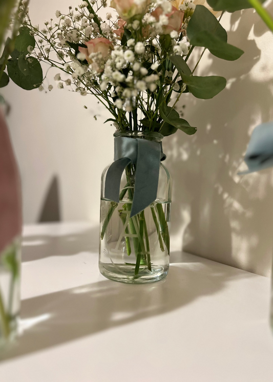 Personalised apothecary-shaped flower vase with the letter A engraved in a delicate oval border, tied with a blue sage green velvet ribbon, and filled with gypsophila, eucalyptus, and soft pink roses, perfect for showcasing floral arrangements and engraved vases for sale.