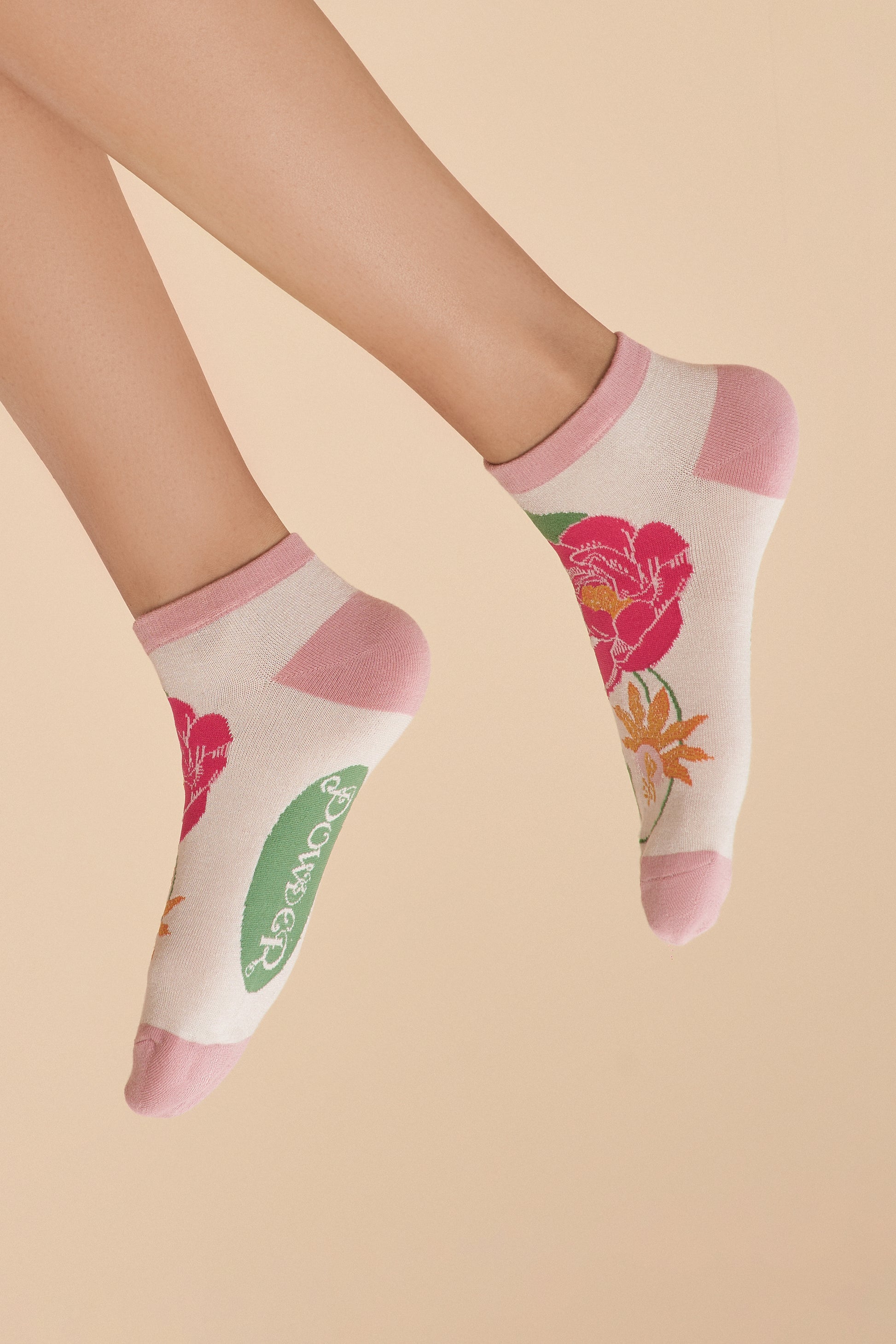 Two feet wearing Tropical Flora Trainer Socks in a coconut-coloured base with pale pink toes and heels, featuring a bright pink and orange floral pattern, a vibrant gift option available from The Petal and Plant Company